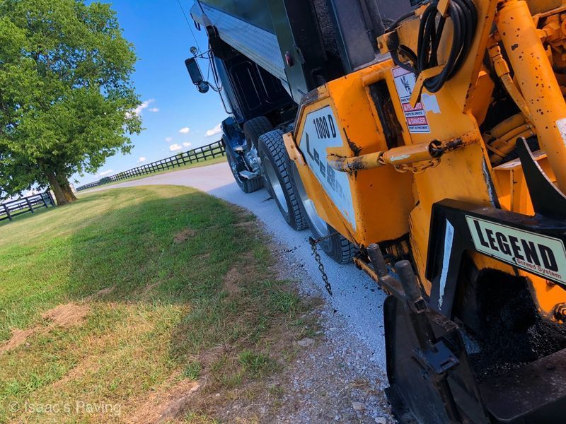 A yellow Legend asphalt paver sits on a gravel road next to a black dump truck with a large tree in a grassy field.