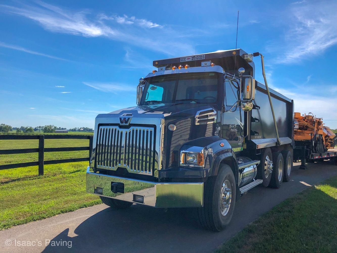 A black Western Star dump truck parked on a paved road with a sunny blue sky background and a grassy field nearby.