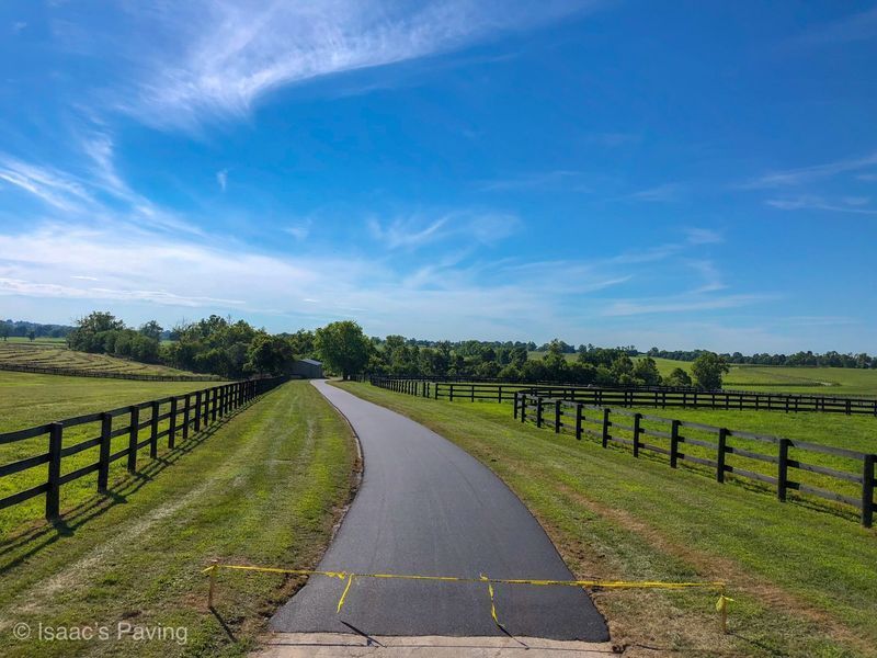 A newly paved asphalt path curves through a grassy field, bordered by black wooden fences under a clear blue sky.