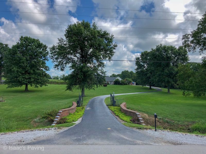 A paved driveway leads through a brick entrance gate into a grassy field with large trees under a blue, cloudy sky.