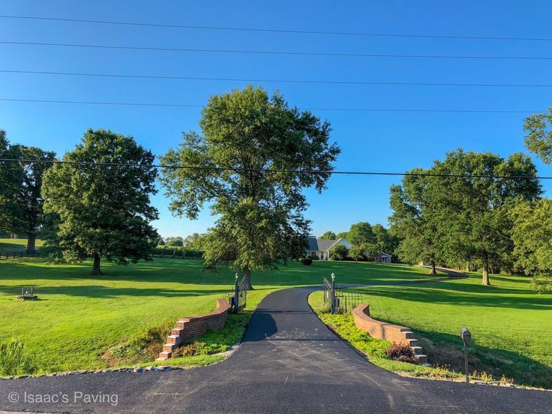 A paved driveway leads through a brick-walled entrance into a green, tree-filled landscape under a clear blue sky.