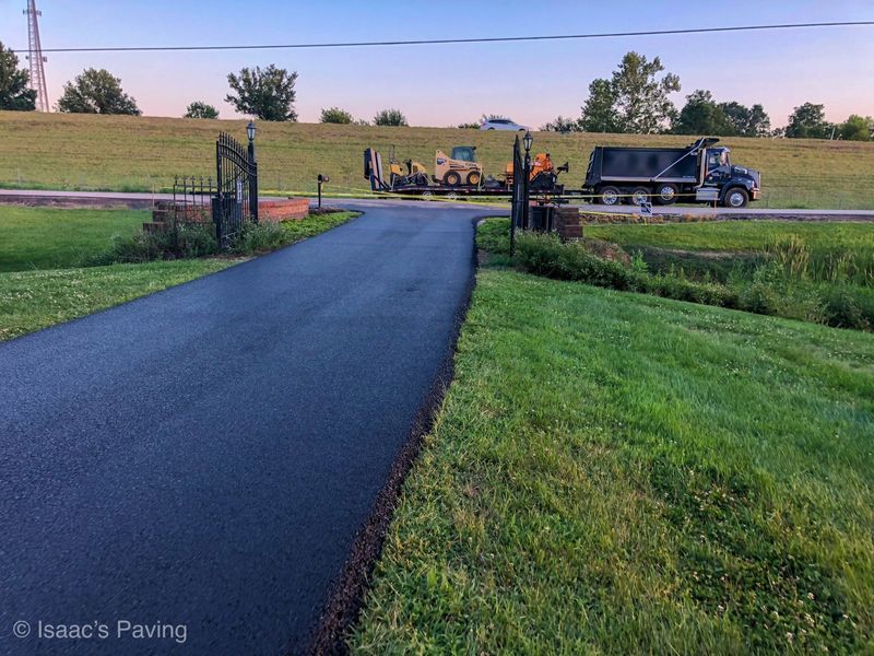 A freshly paved black driveway leading to an open iron gate, with construction equipment parked on the road beyond.