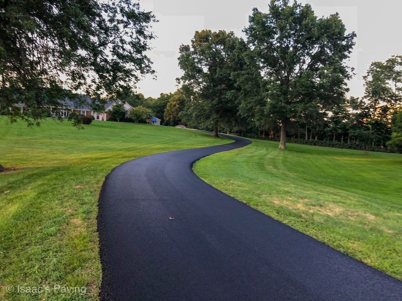 A curved black asphalt driveway winds through a lush green lawn toward a house surrounded by trees.