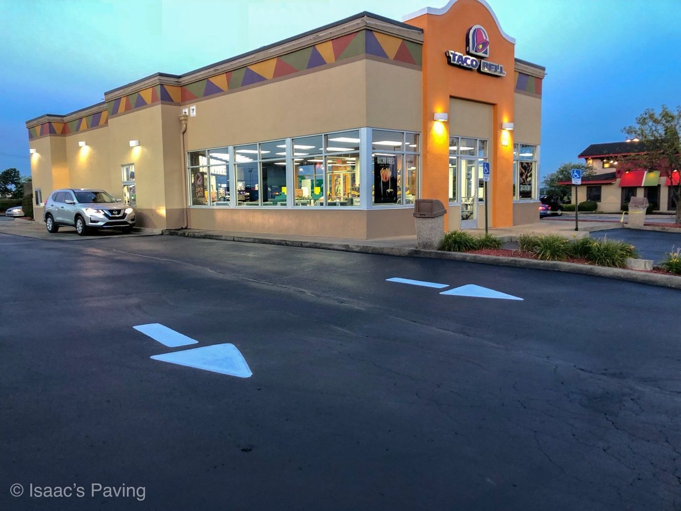 A tan Taco Bell restaurant at dusk with two white directional arrows painted on the asphalt parking lot.