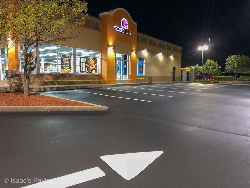 A Taco Bell restaurant exterior at night with a paved parking lot and a large white directional arrow on the ground.