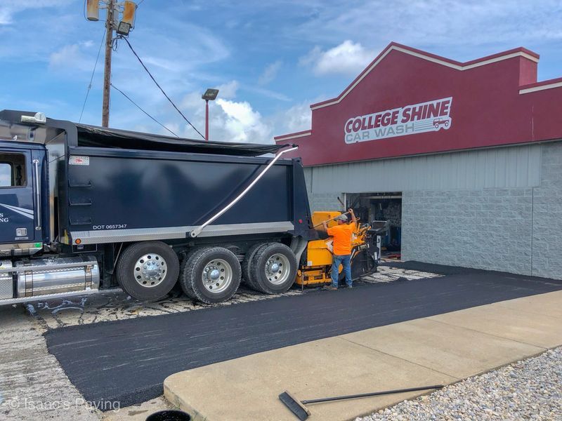 A worker in an orange shirt operates a paving machine to lay asphalt in front of a College Shine car wash building.