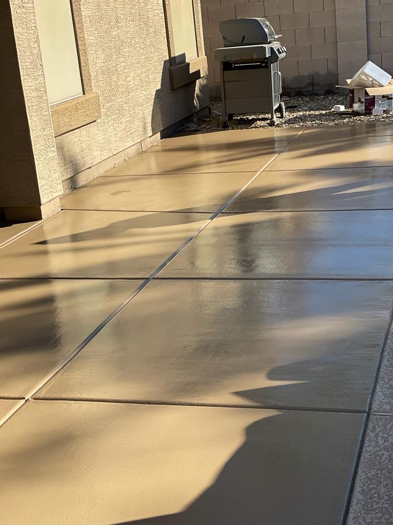 Shiny, tan concrete patio with rectangular sections. A grill and white object sit in the background near a stucco wall.