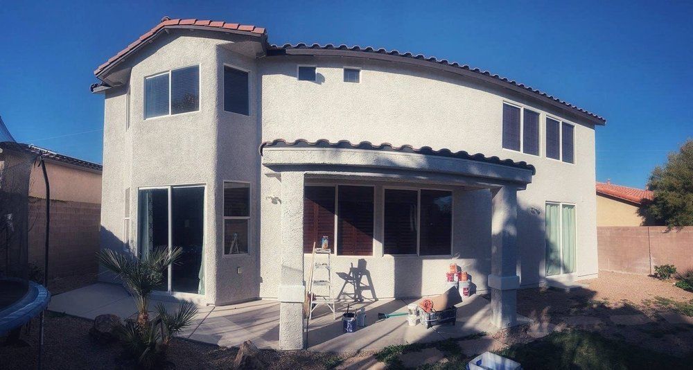 Two-story stucco house with curved back, tiled roof, windows, and a covered patio under a bright blue sky.