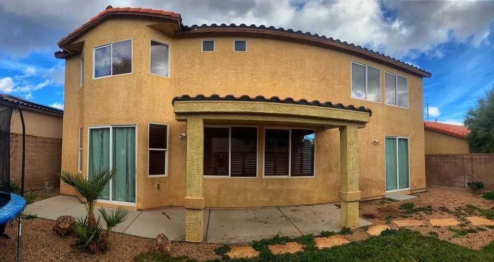 Back exterior of a two-story stucco house with a tiled roof and covered patio, under a blue sky.