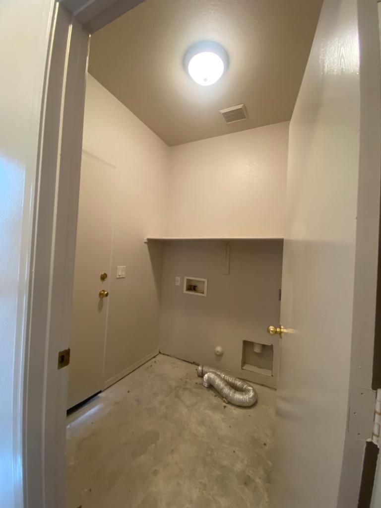 Laundry room with bare concrete floor, beige walls, and a shelf above washer/dryer connections.