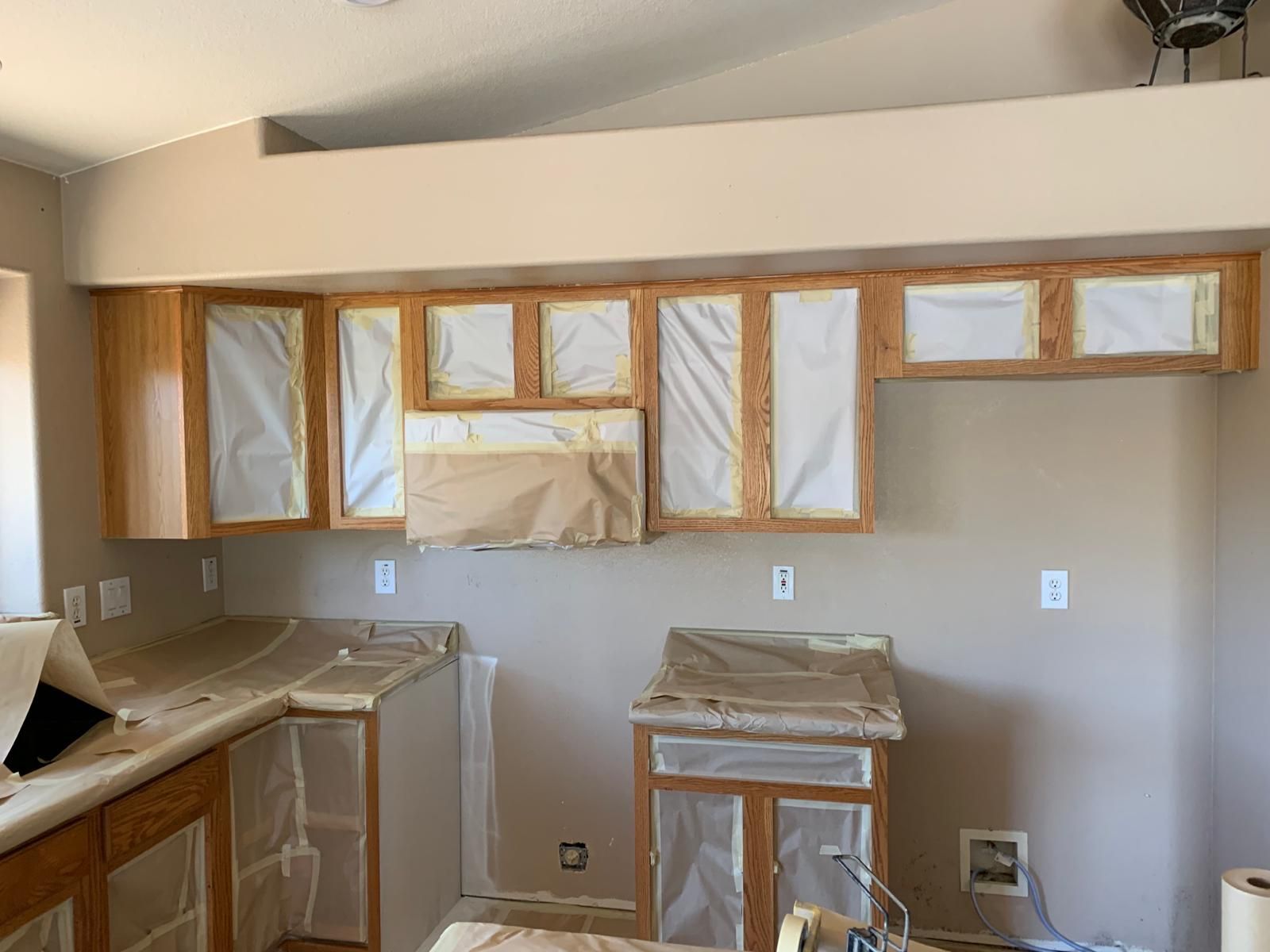 Kitchen cabinets masked with paper, ready for painting. Light wood cabinets and countertops. Beige walls.