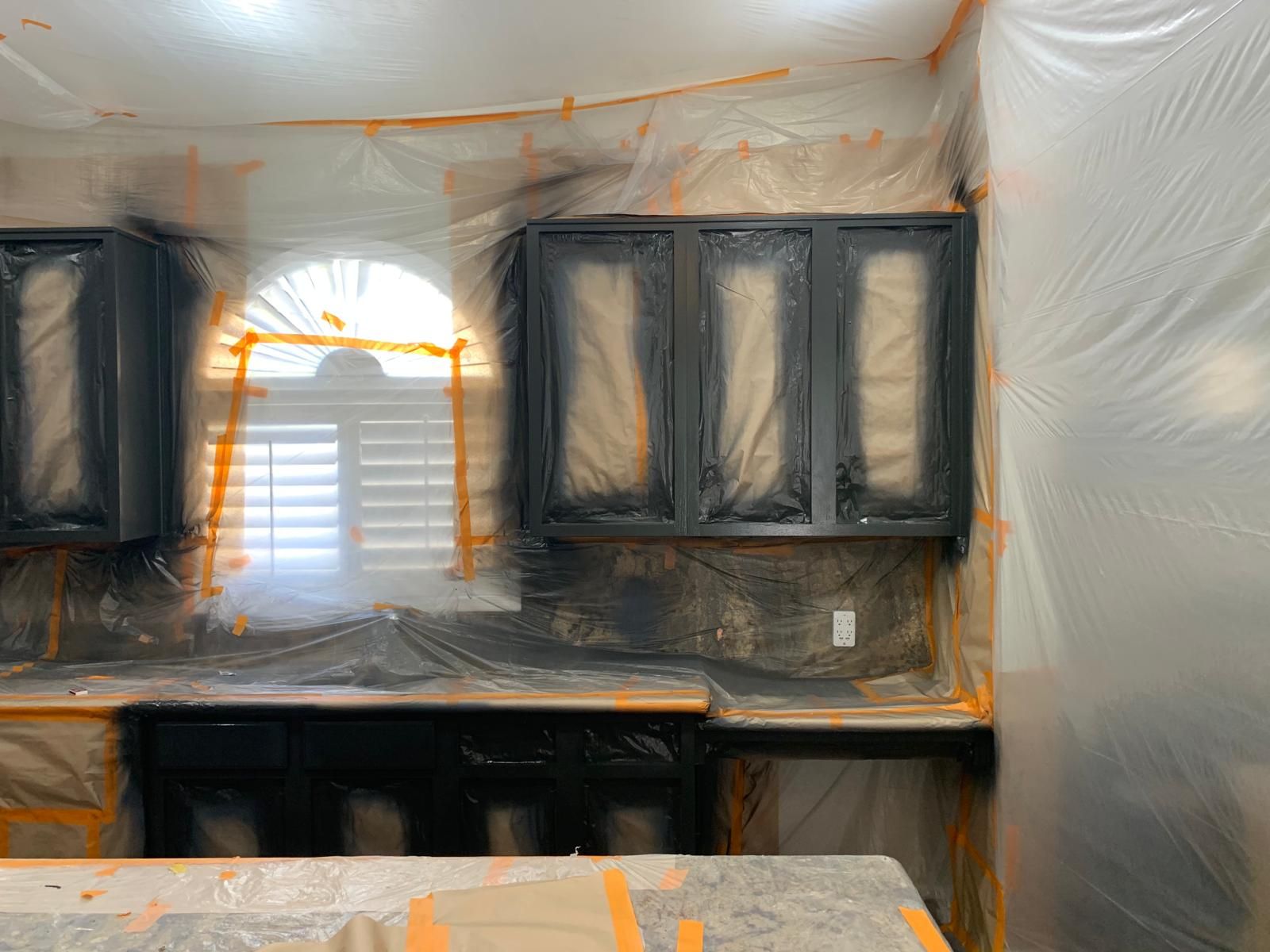 Kitchen cabinets being spray-painted black, covered in plastic and masking tape. Window is shuttered.