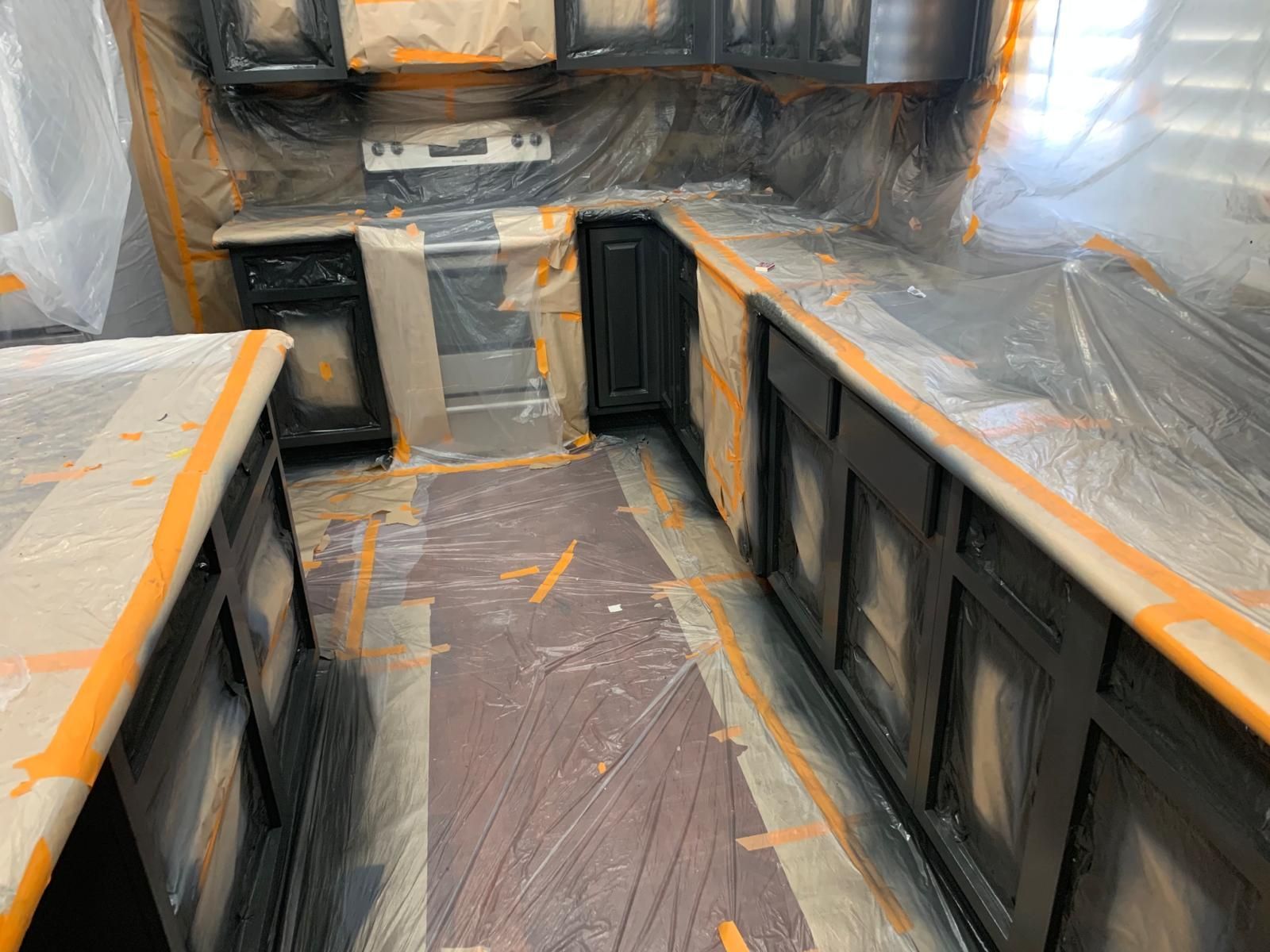 Kitchen cabinets being painted black, protected by tape and plastic sheeting.