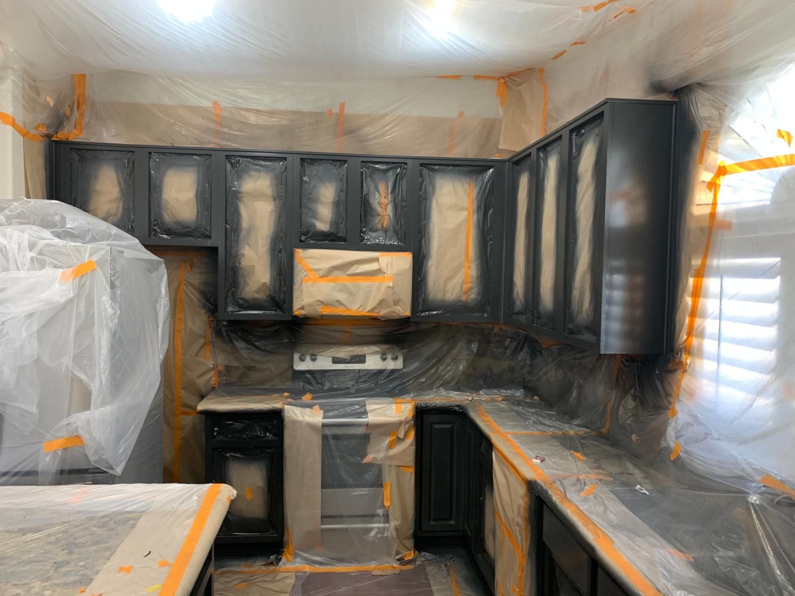 Kitchen cabinets being painted dark grey, with surfaces covered in plastic for protection.