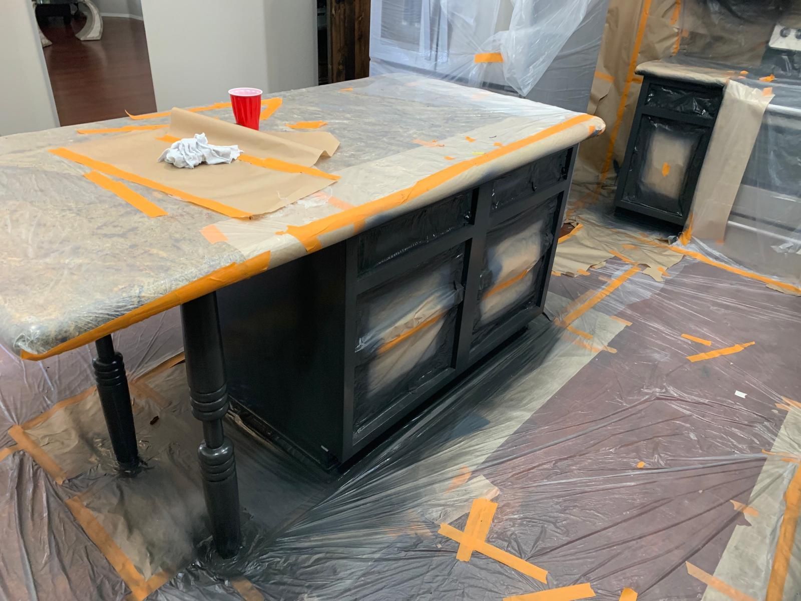 Kitchen island being painted black, protected by tape and plastic. A cup and cloth rest on the countertop.