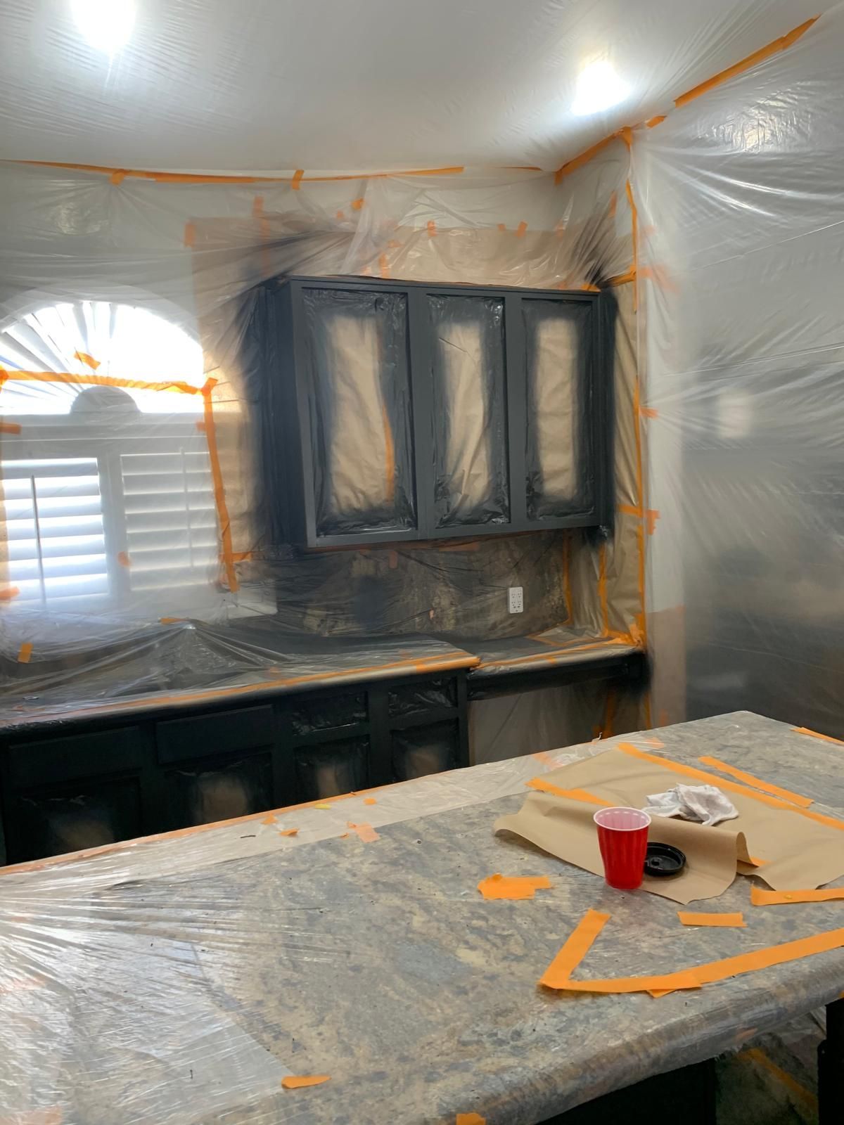 Kitchen cabinets being painted black; interior partially covered with plastic and taped for protection.