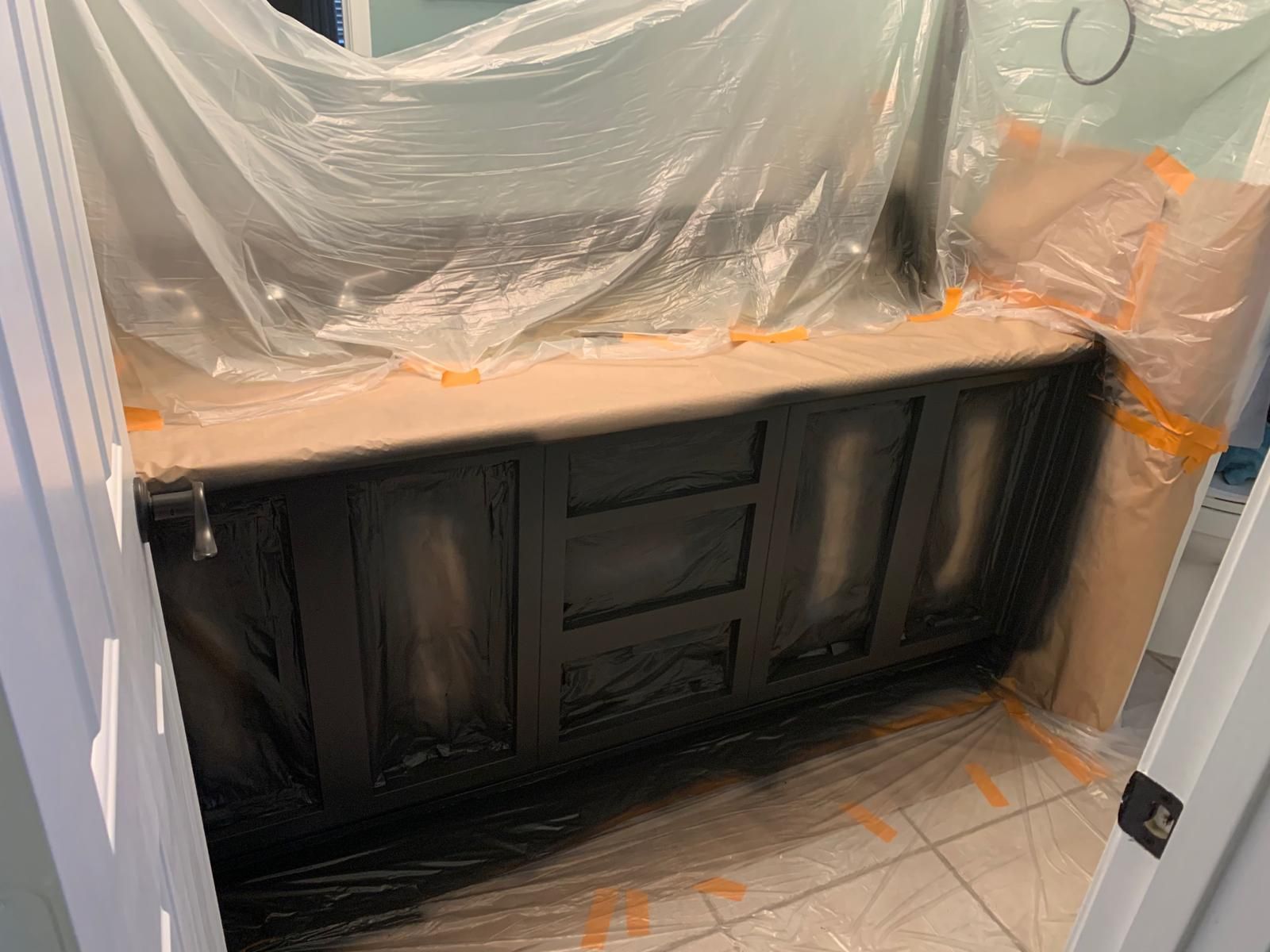 Vanity cabinet being painted black, protected by plastic sheeting in a bathroom.