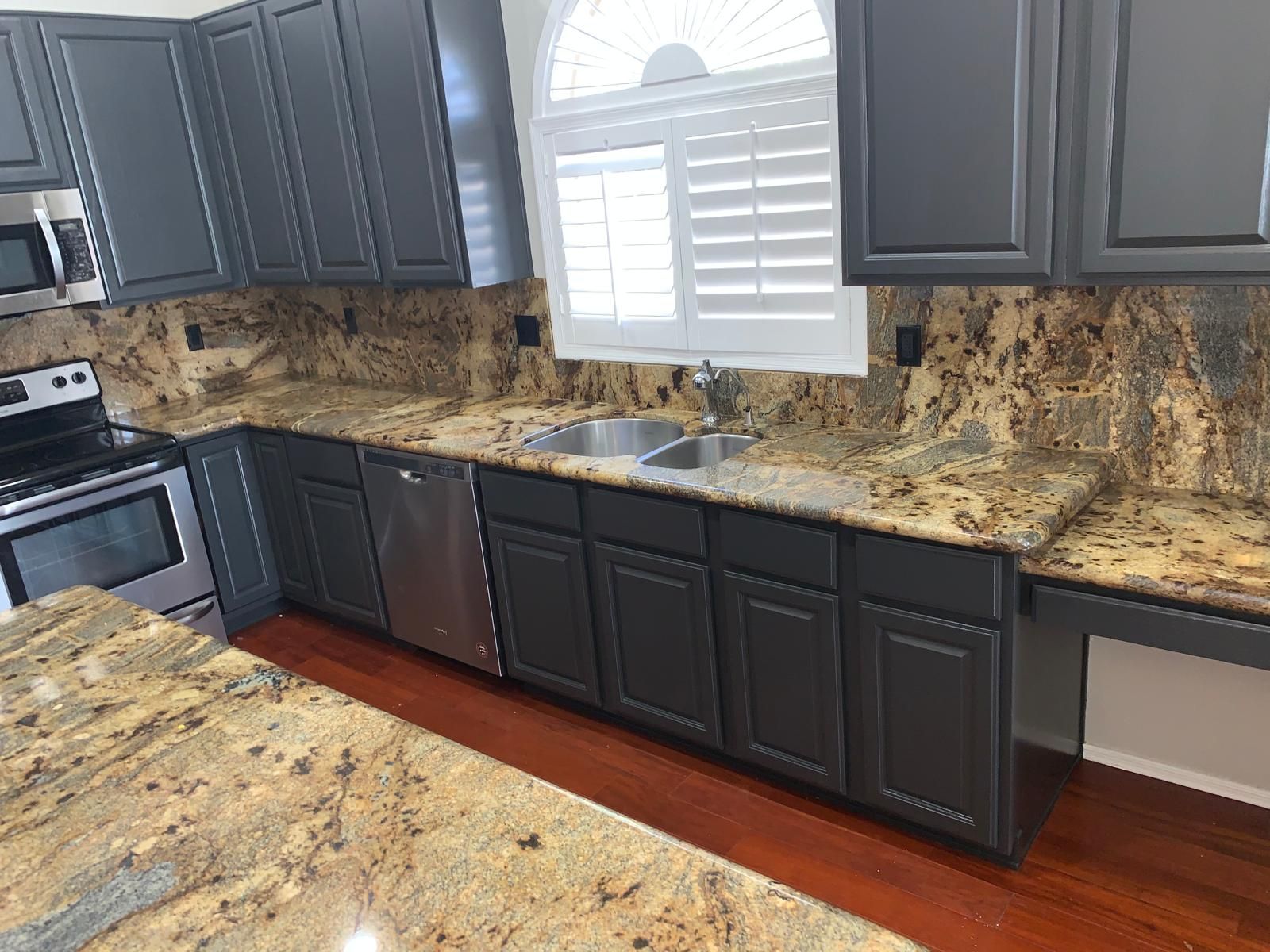 Dark gray kitchen cabinets with a granite countertop, stainless steel appliances, and a window with white shutters.