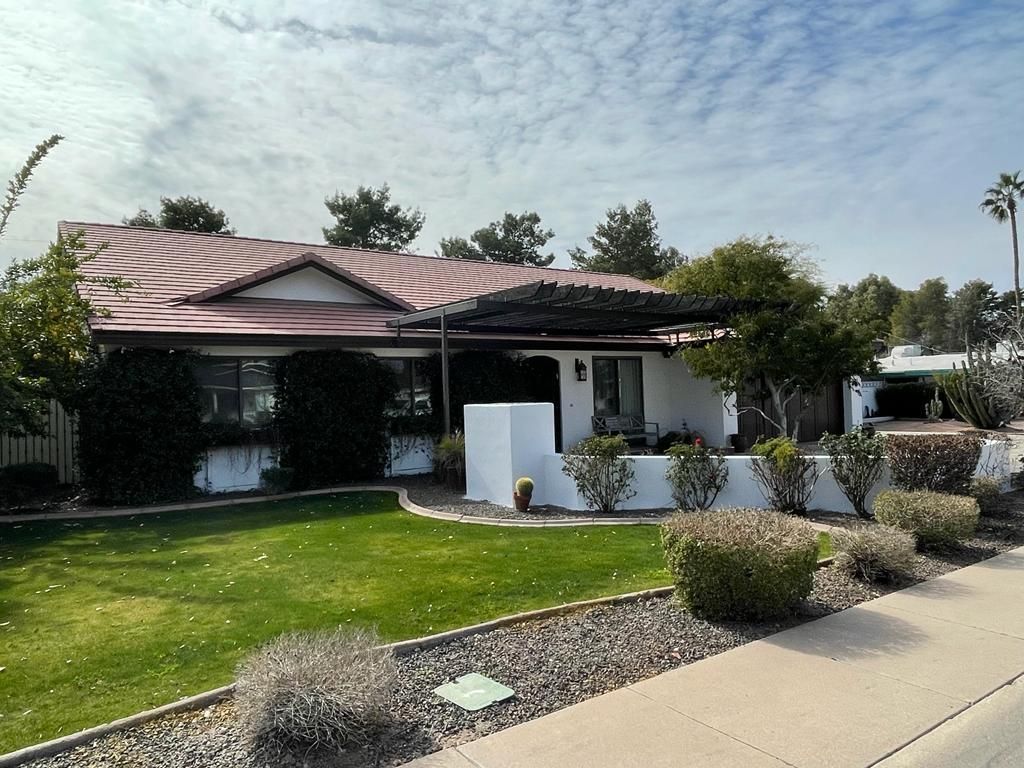 A one-story white house with a brown tile roof, green lawn, bushes, and a covered patio.