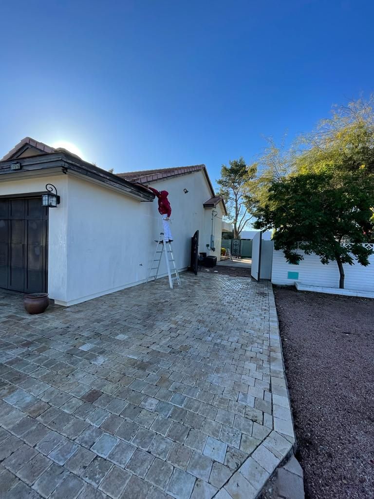 Person on a ladder painting a white stucco house on a sunny day. A brick patio and small trees are in view.