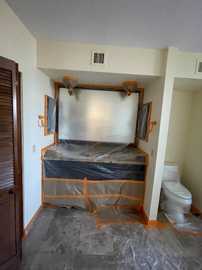 A bed is covered with plastic and tape, possibly being painted. A toilet is visible in the adjacent room.