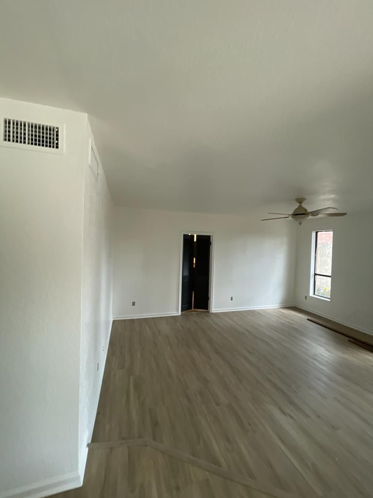 Empty, white-walled room with wood-look flooring. A doorway and window are visible. A ceiling fan is present.