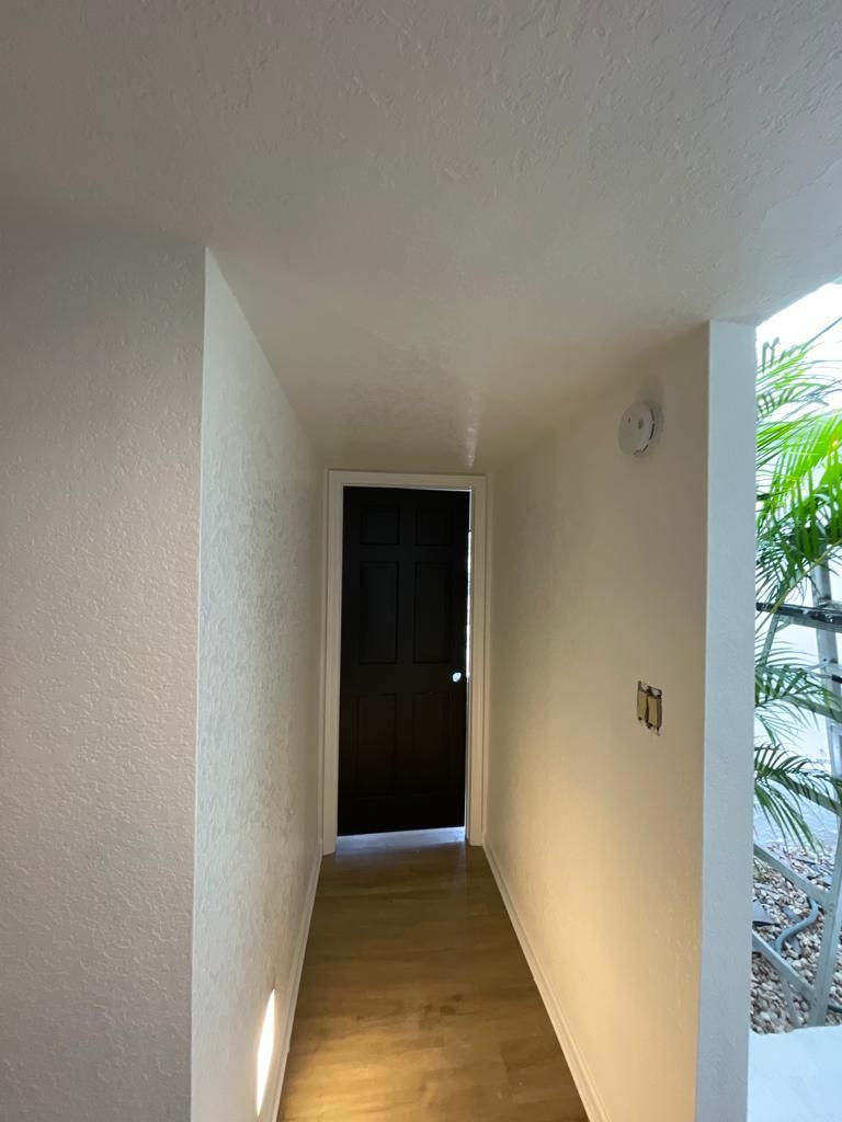 Narrow hallway with light walls, wood floor, and a dark door at the end. Sunlight enters from the right.