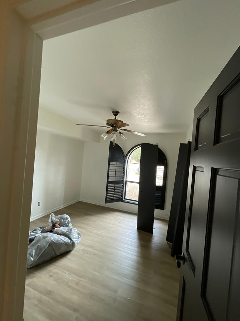 Empty room with arched window, dark shutters, and ceiling fan; gray floor, white walls.