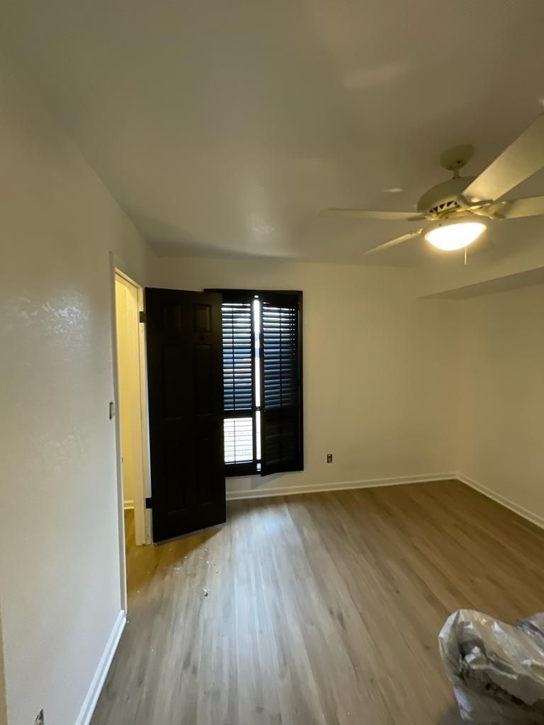Empty room with wood-look flooring, white walls, black shutters, and a ceiling fan. A doorway leads to another room.