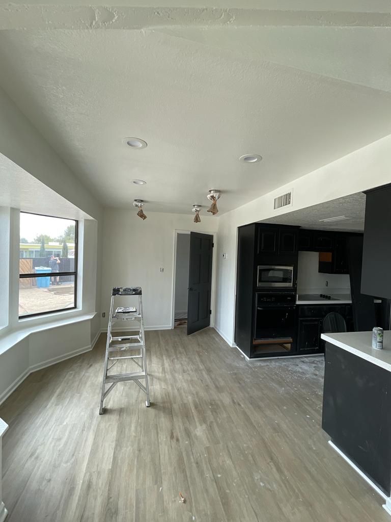 Interior room with a bay window, kitchen, ladder, and black cabinets. Light wood flooring.