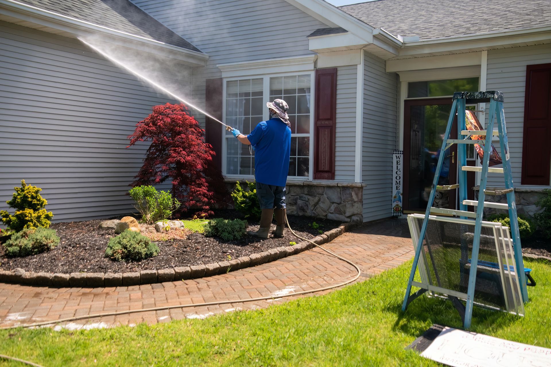 Person power washing a house with a blue hose, near a red shrub, ladder, and brick walkway.