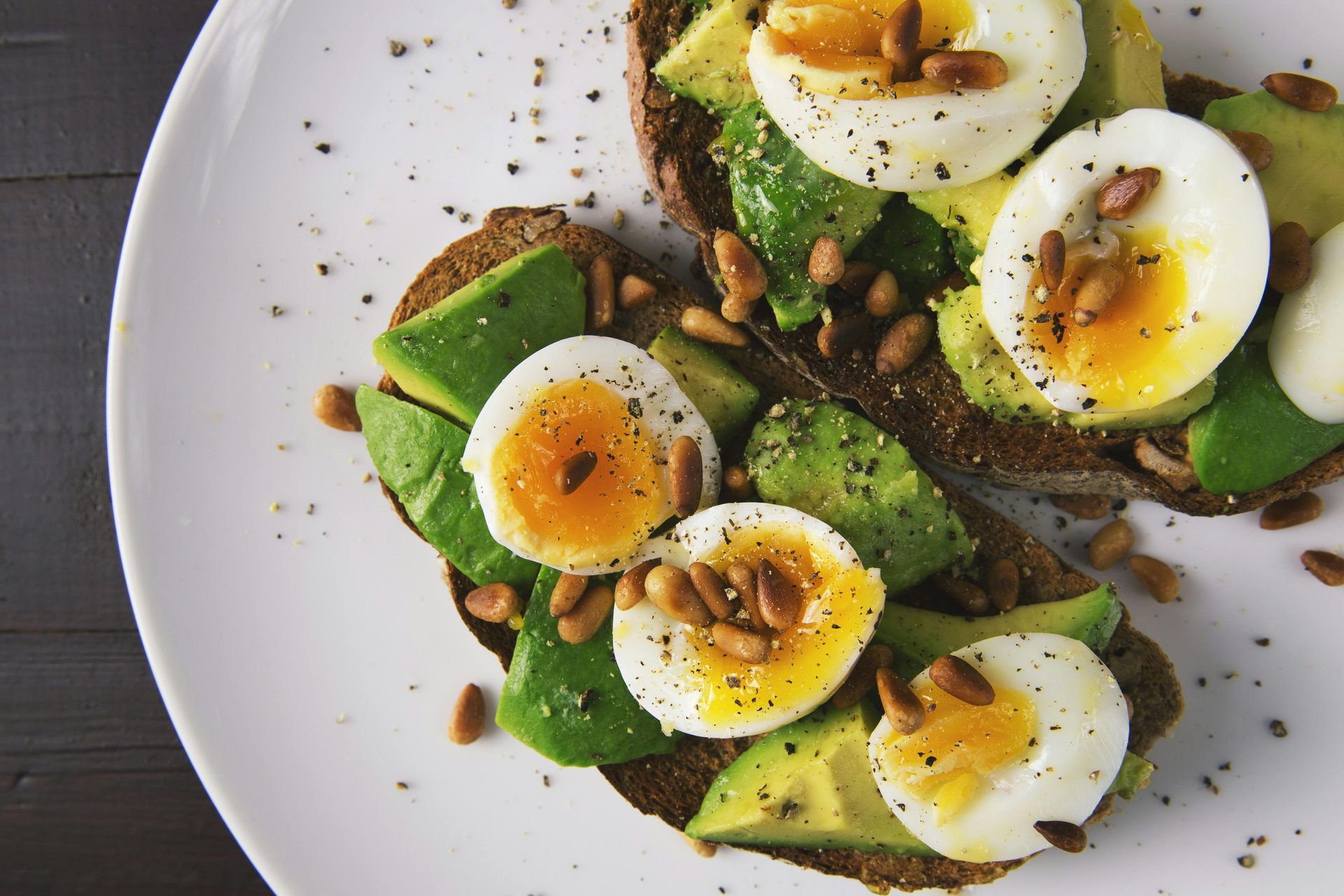 Two slices of toast with avocado , hard boiled eggs and pine nuts on a white plate.