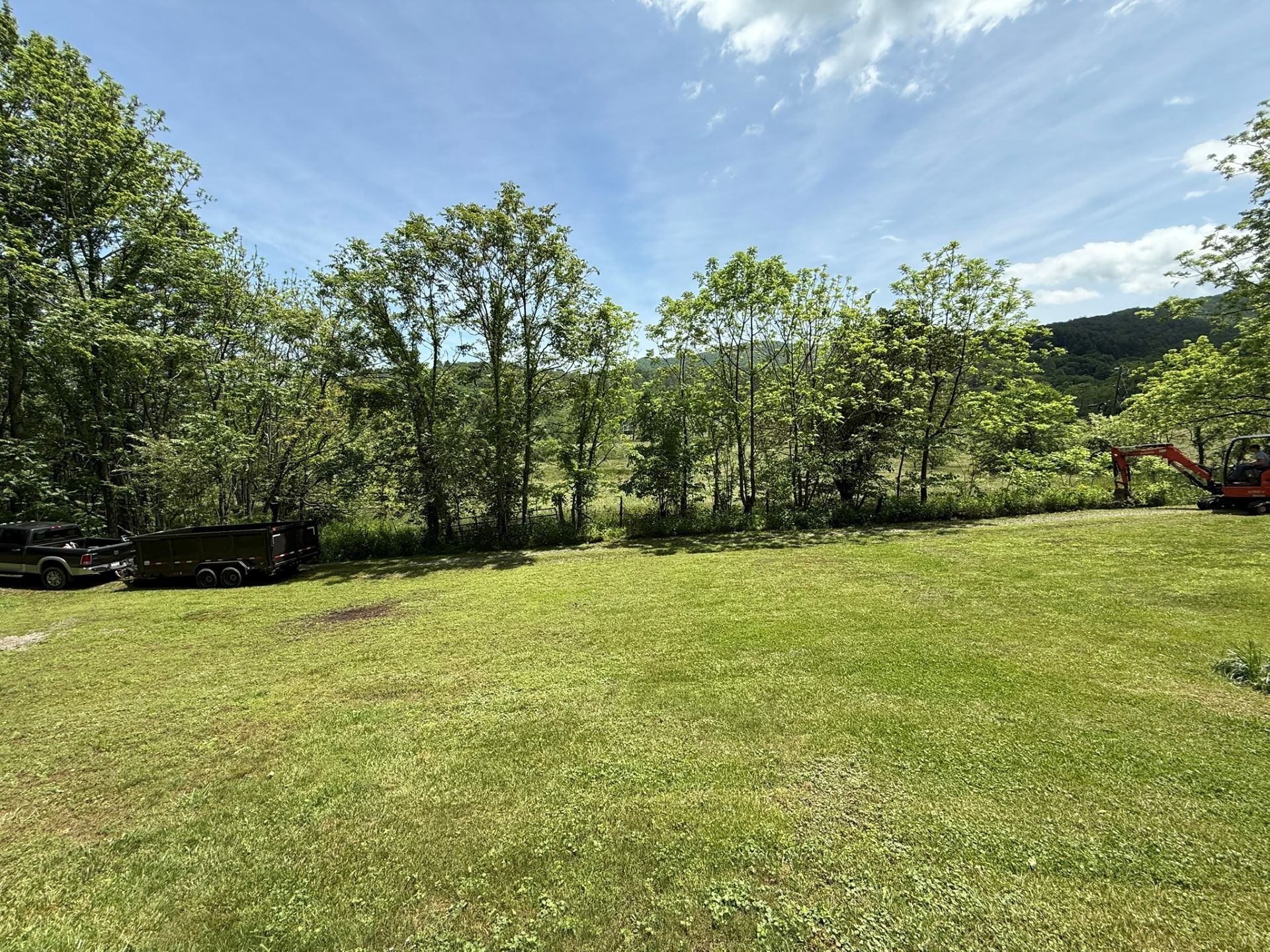 Grassy field with trees, blue sky and a construction vehicle in the distance.