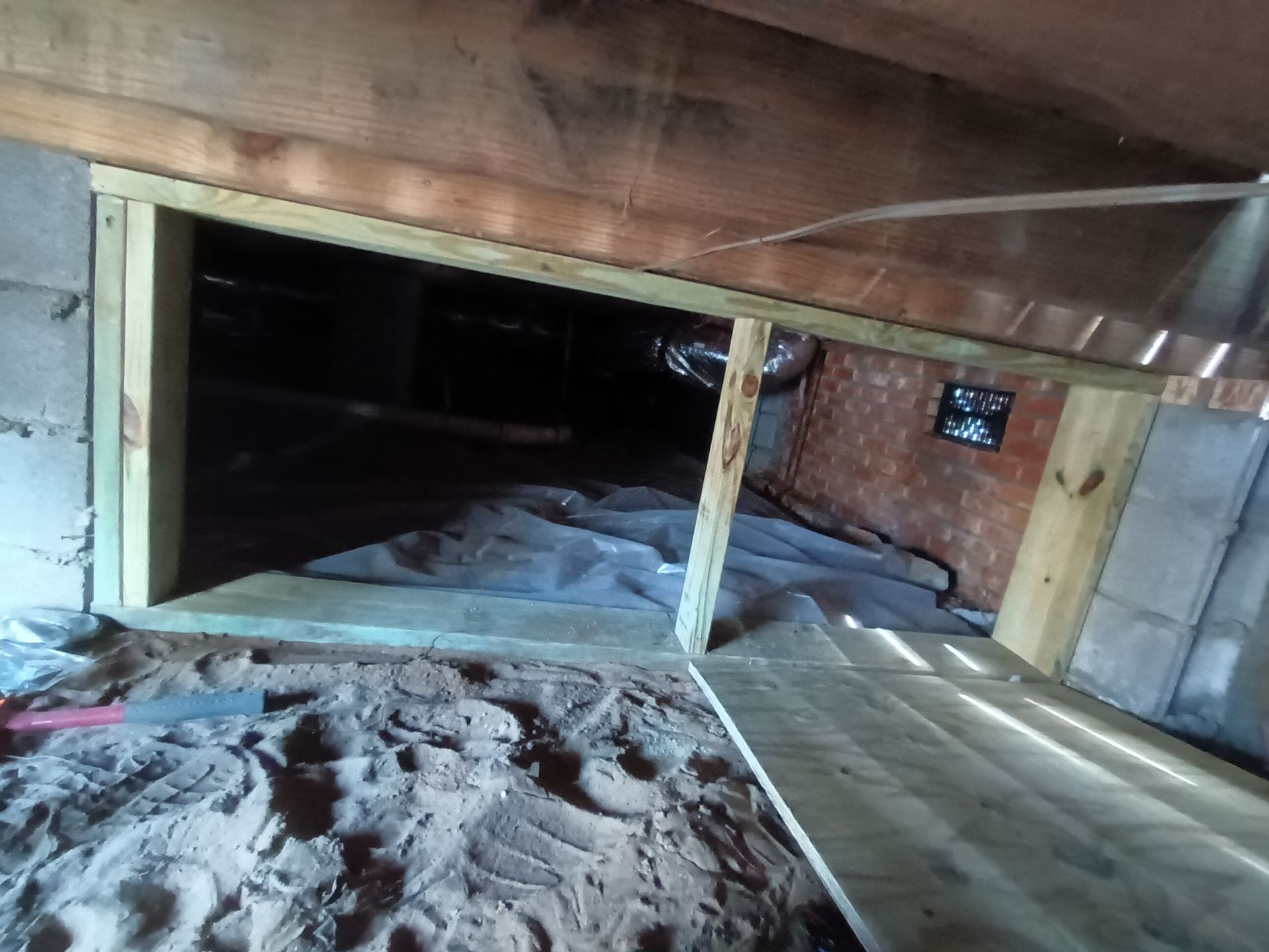A crawlspace opening framed with pressure-treated lumber, showing sandy ground and a plastic vapor barrier underneath.