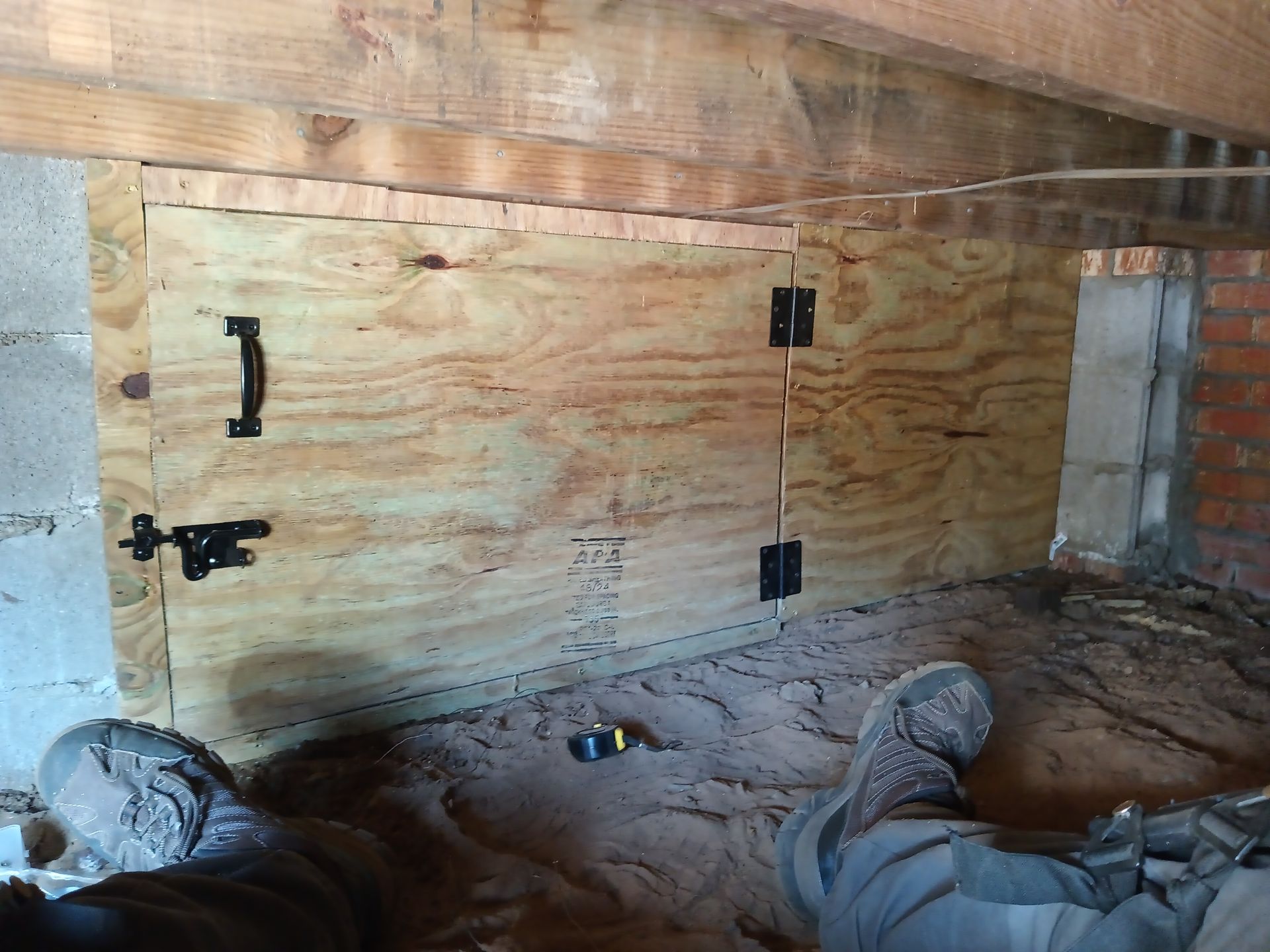 A plywood access door with a handle and latch installed in a crawl space between a concrete and brick foundation.