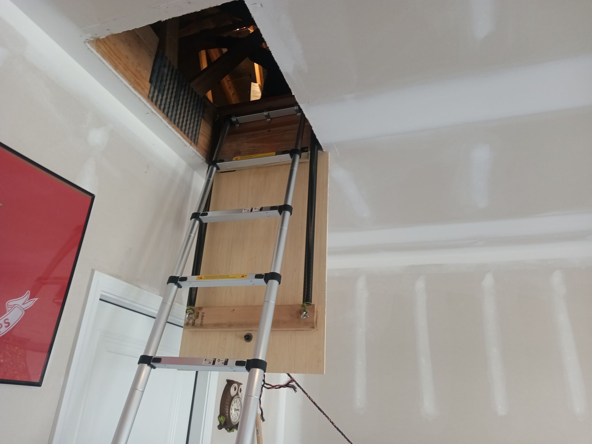 An aluminum telescoping ladder is extended through a rectangular ceiling hatch into an attic space.