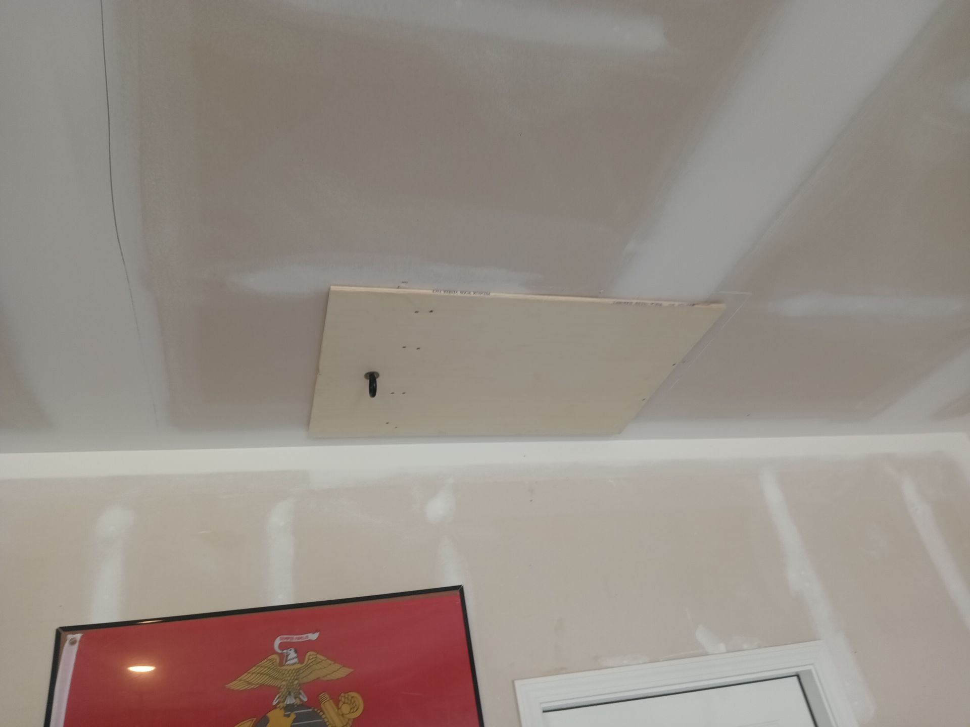 A rectangular attic access panel is installed in a drywall ceiling, positioned above a wall with a framed red emblem.