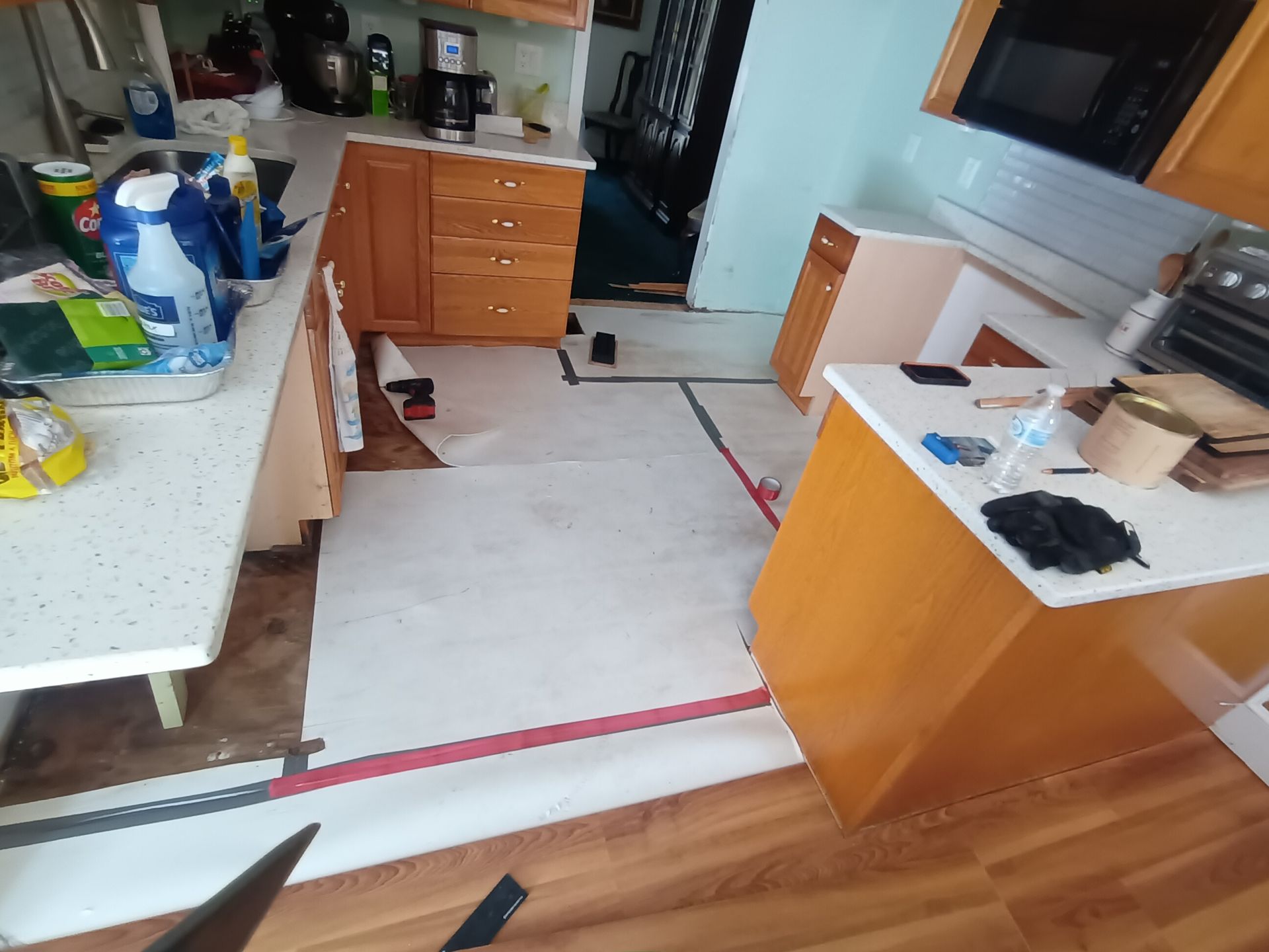 A kitchen under renovation with wood cabinets, exposed subflooring, and protective paper laid over sections of the floor.