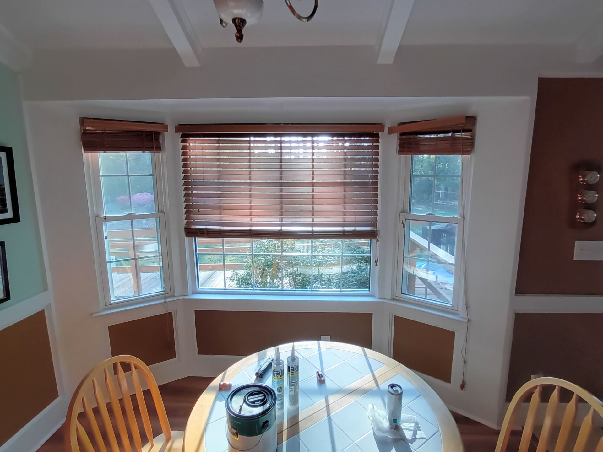 A dining nook with a white round table, two wooden chairs, and a bay window featuring brown wood blinds.