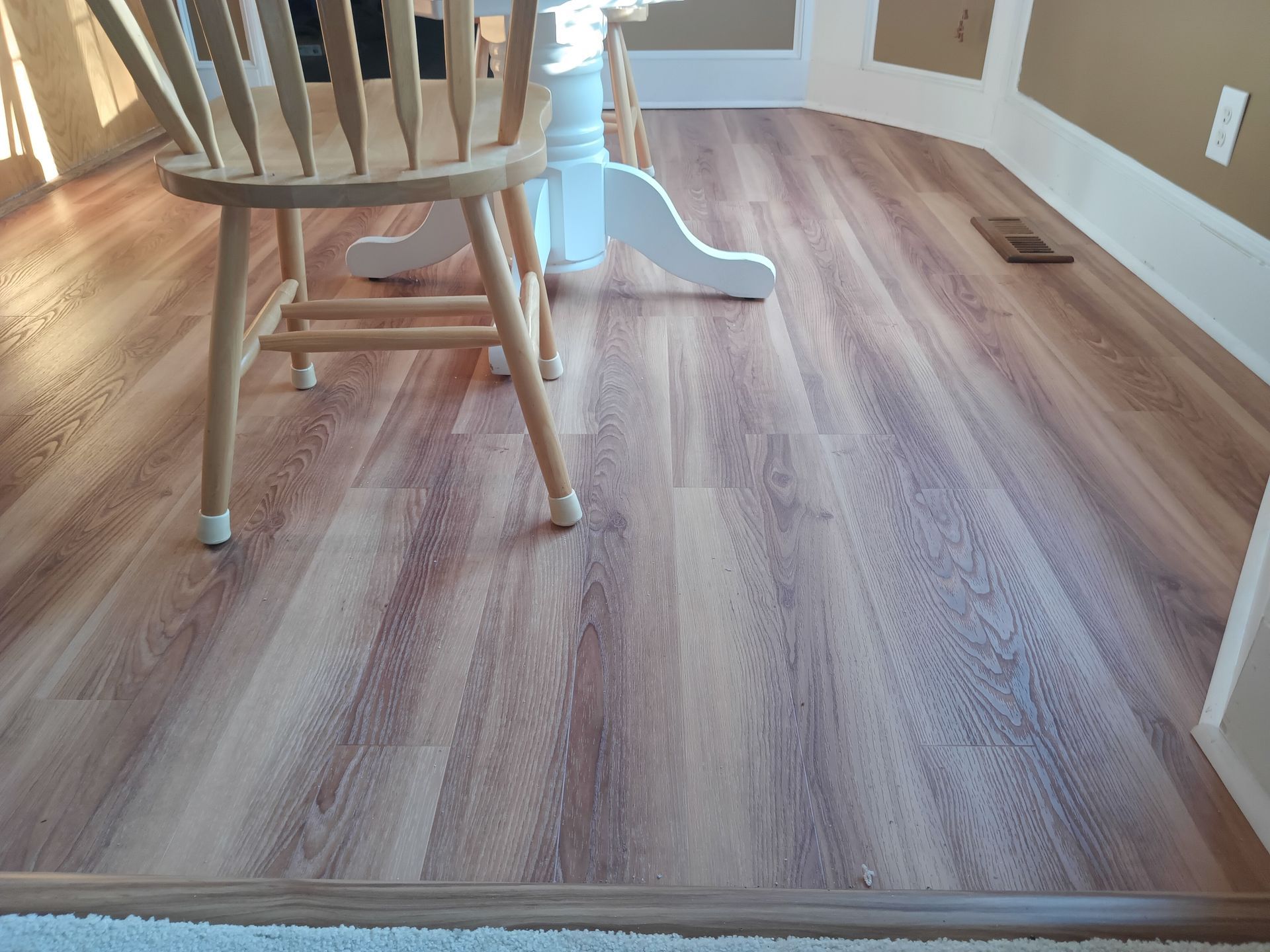 A wooden chair stands on light brown wood-look flooring next to a white table base in a room with tan walls.