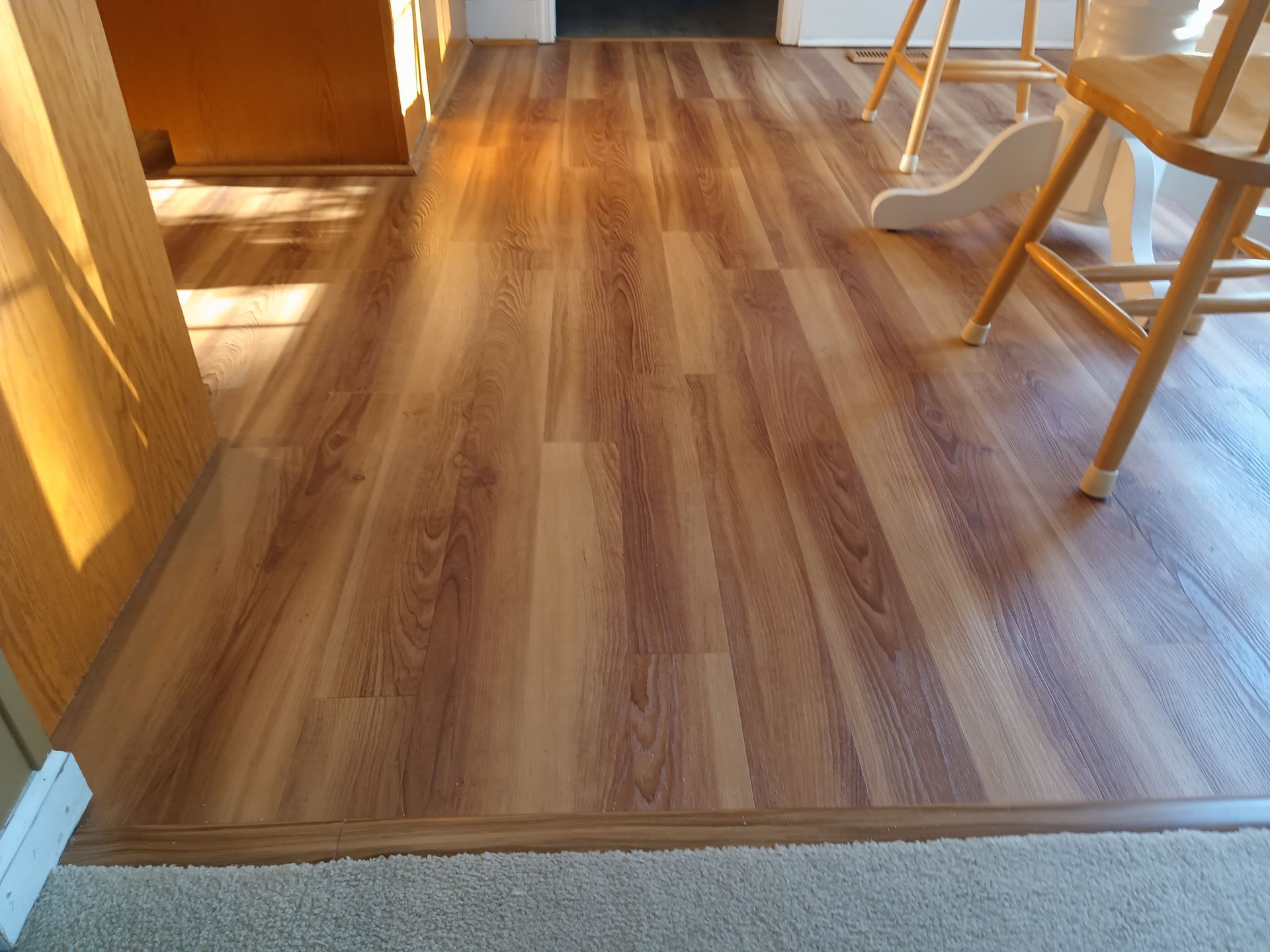 A hardwood-style vinyl floor meets a carpeted area, with wooden chairs visible in the background.