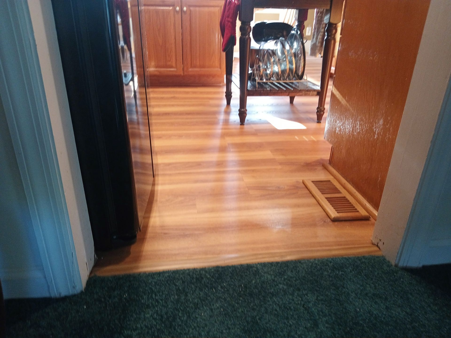 A view from a carpeted room into a kitchen with wood laminate flooring, a wooden table, and a floor vent.