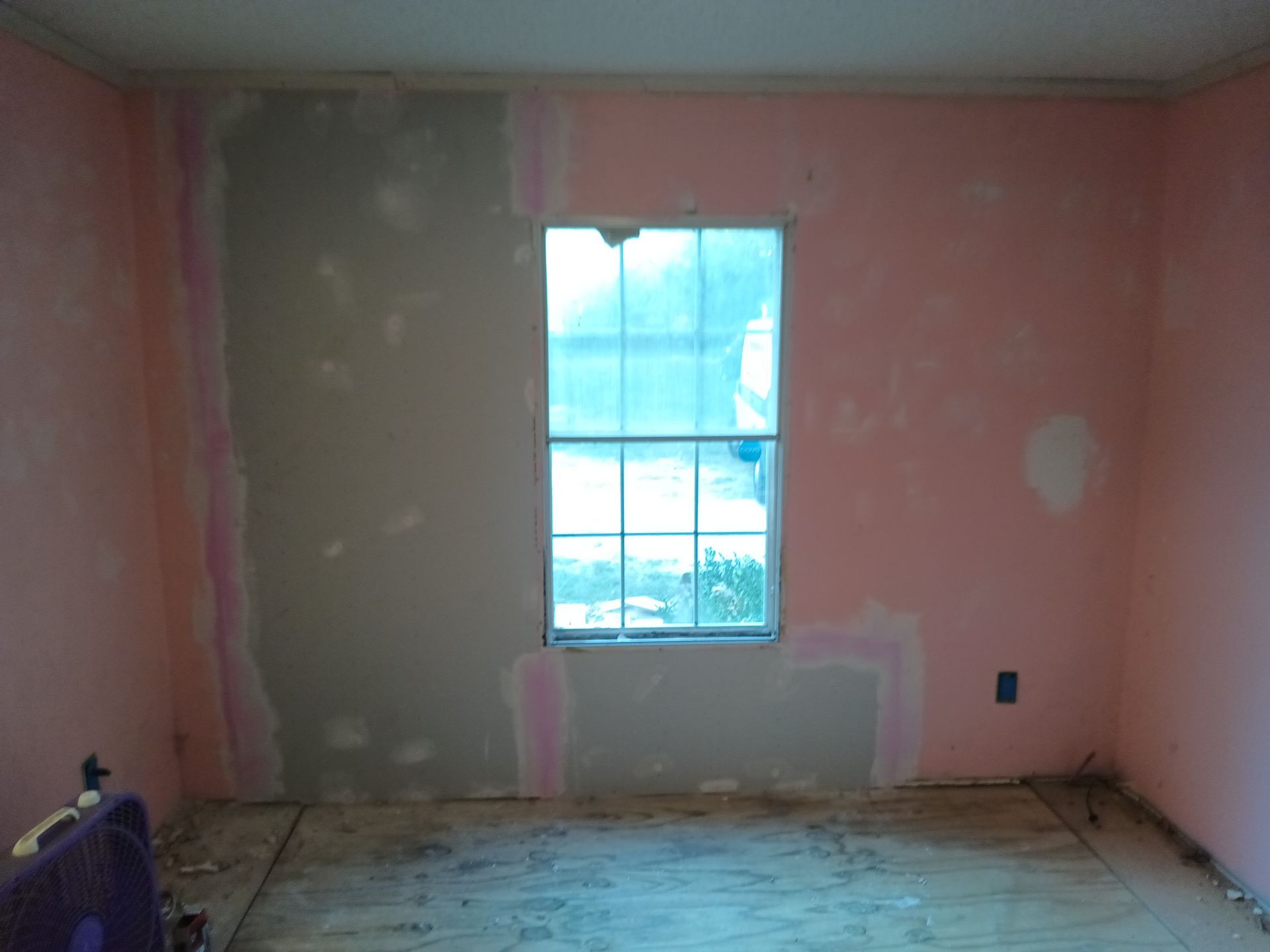 A room undergoing wall repairs, featuring a window and patches of pink paint and grey joint compound on the walls.