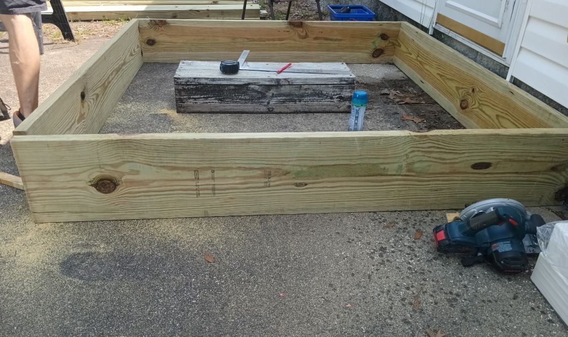 A partially assembled wooden rectangular frame for a raised garden bed sits on a gravel surface.