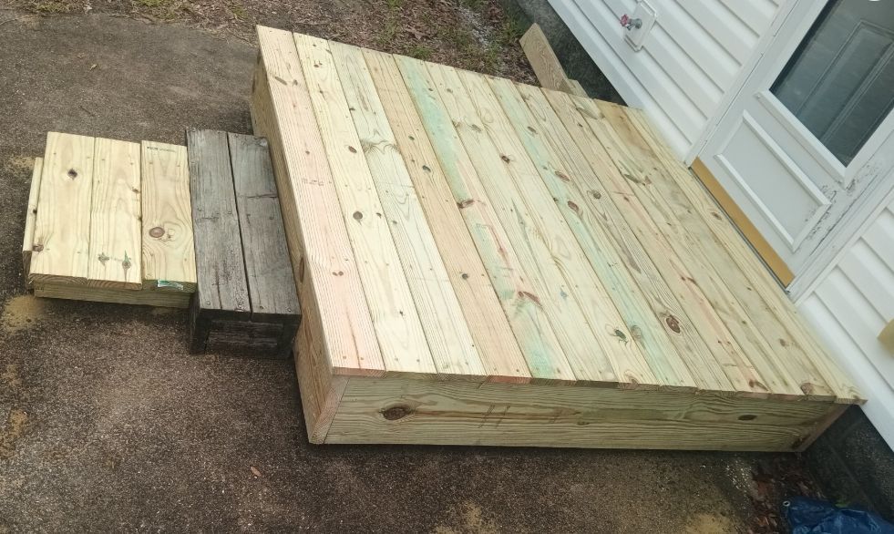 A small, newly constructed wooden deck platform outside a door, with an uneven temporary step made of boards and a log.