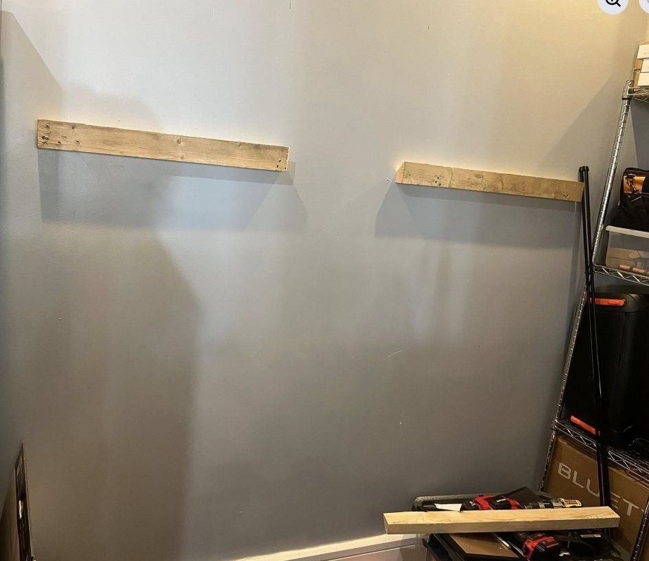 Two wooden boards are mounted horizontally on a light gray wall, with an additional board resting on the floor below.