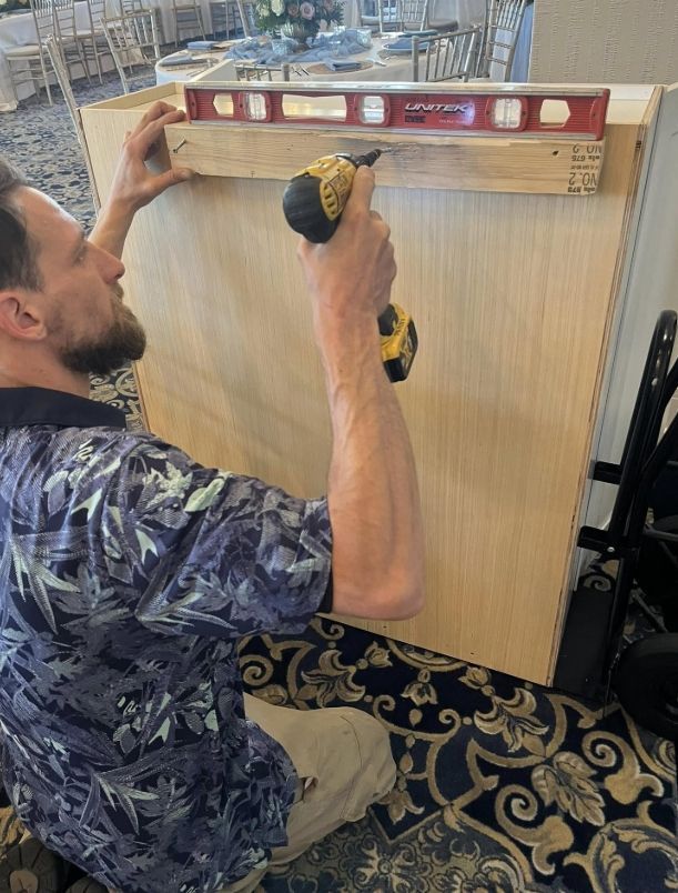 A person uses a level and a cordless drill to attach a wooden support rail to a cabinet panel in an indoor setting.