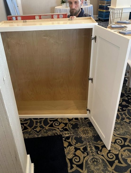 A person installs a white, open-faced wooden cabinet on top of a patterned carpet in a room with light-colored walls.
