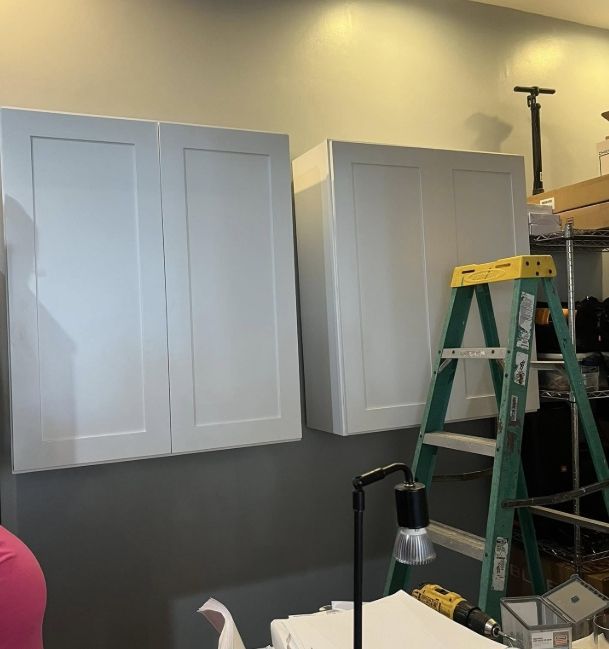 Two white kitchen cabinets are mounted on a gray wall next to a green step ladder and a small desk lamp.