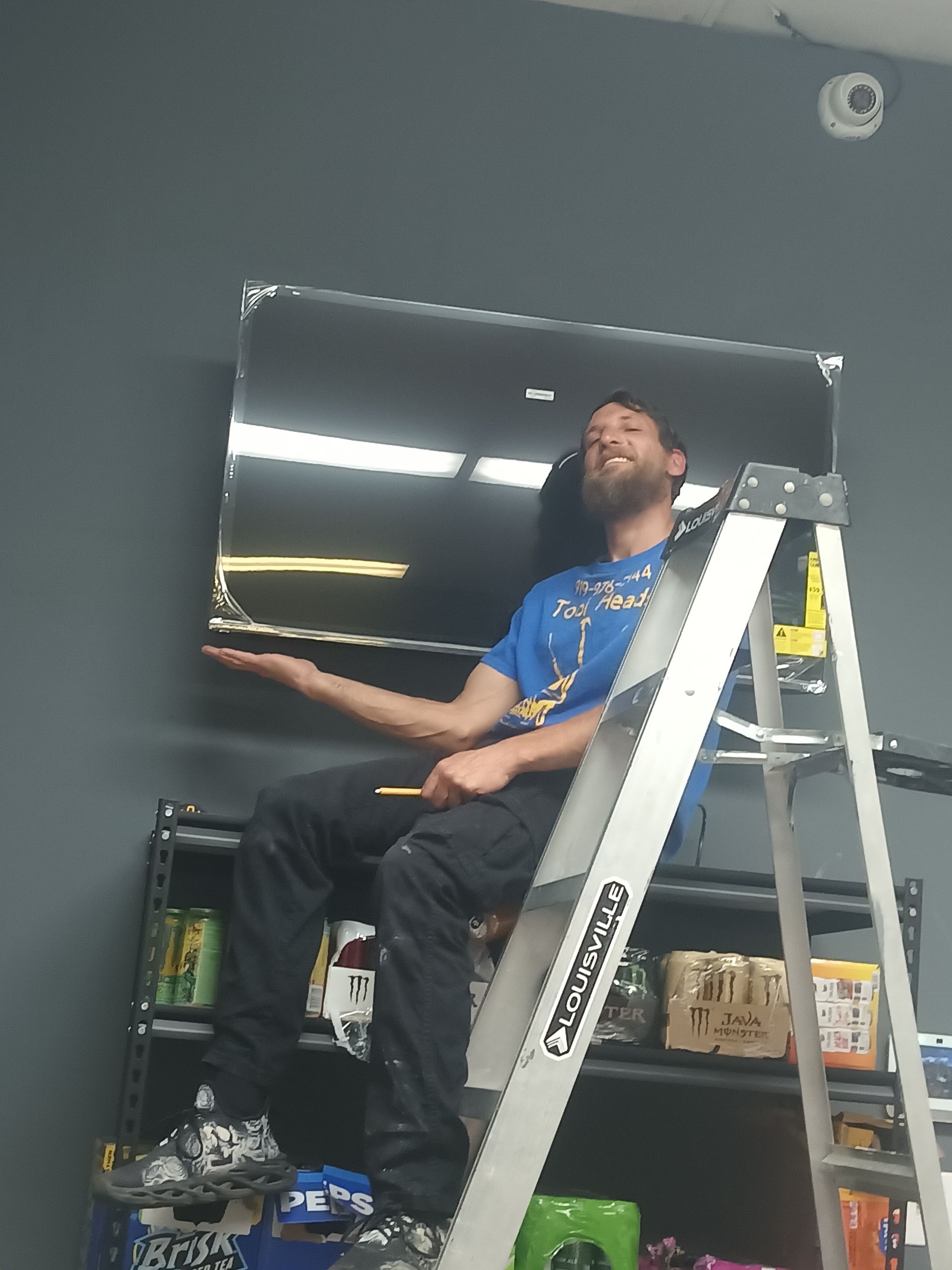 A person sits on a ladder in front of a gray wall, smiling while holding up a large, wall-mounted television.